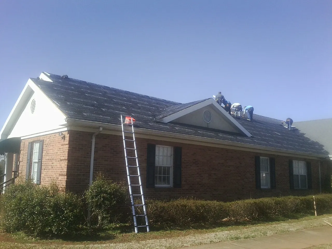 Crew tearing shingles off a brick ranch roof with a ladder set — Emergency Roof Repair work by our crew in Mandan