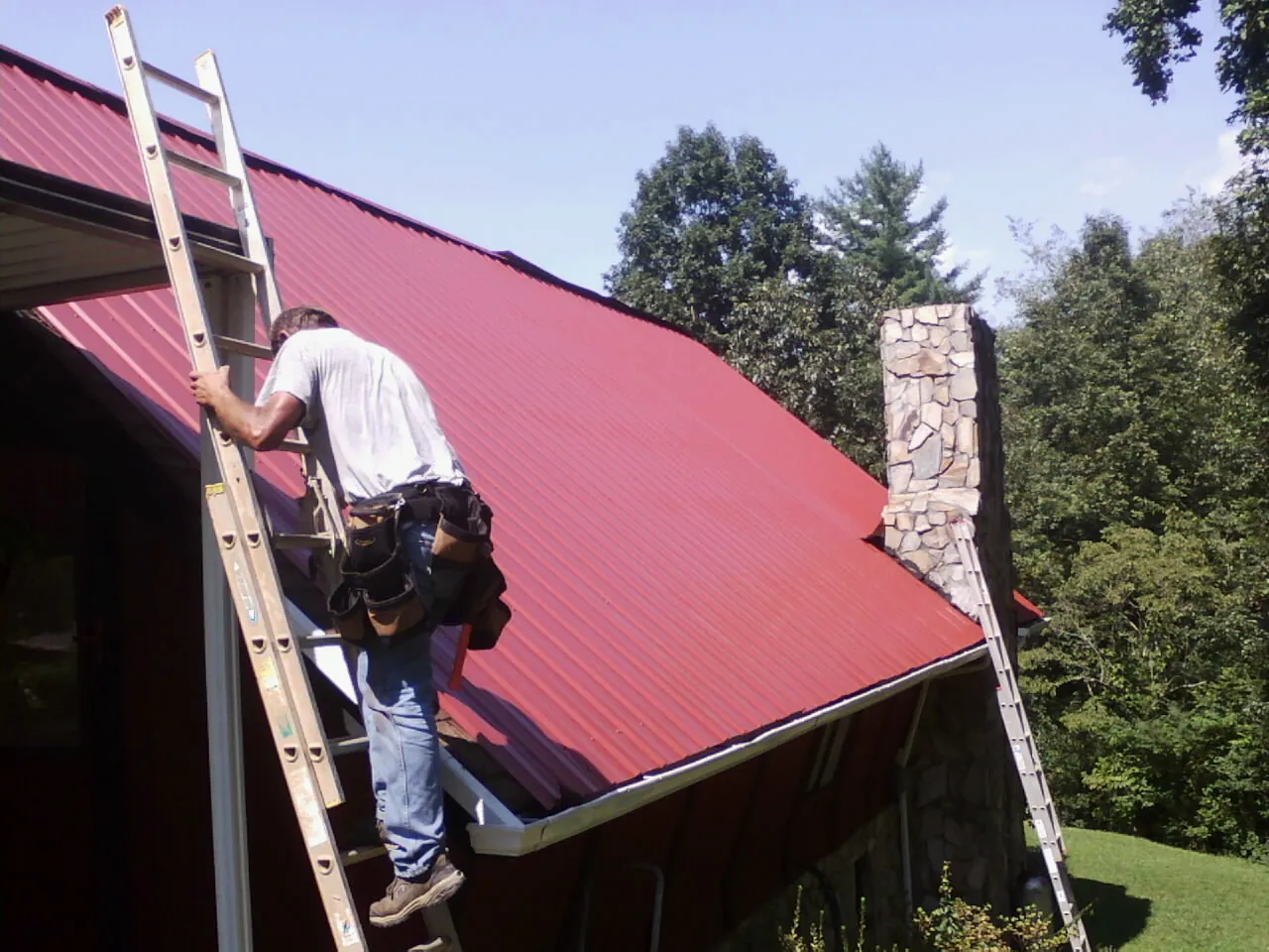 Roofer on a ladder beside a red metal roof and stone chimney — Soffit & Fascia Repair by Roofing Brothers in Mandan